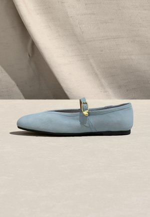 Hellblauer Wildleder-Mary-Jane-Flachschuh mit dünner goldener Schnalle und herzförmigem Anhänger auf neutralem Stoffhintergrund.