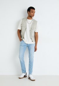 Light grey vest with multiple pockets over a white t-shirt, paired with light blue jeans and white sneakers with black accents.