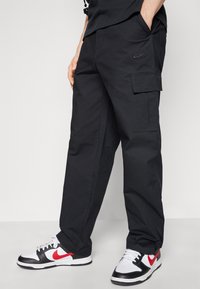 Black cargo pants with a textured finish, side pockets, and a subtle embroidered Nike logo on the thigh. Paired with black, white, and red sneakers.