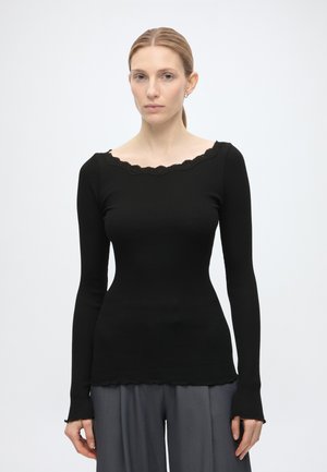 Mujer de piel clara y cabello rubio recogido lleva una camiseta negra ajustada de manga larga con bordes festoneados y pantalones grises holgados, de pie contra un fondo blanco.