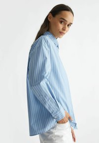 Camisa de manga larga azul claro con rayas verticales blancas, cuello abotonado y un corte relajado. Presenta puños con botones y aberturas laterales.