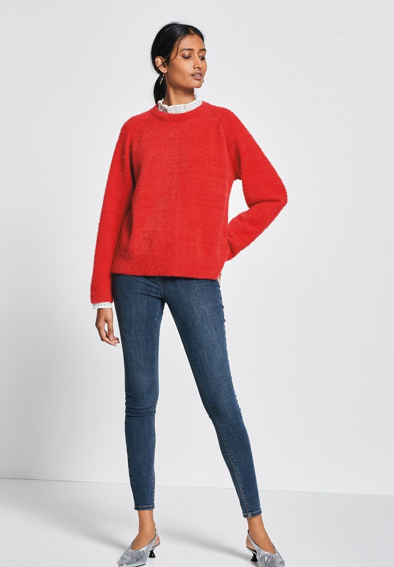 Czerwony puszysty sweter z okrągłym dekoltem i długimi rękawami, nałożony na biały kołnierzyk w falbany. W połączeniu z ciemnymi wąskimi dżinsami i srebrnymi butami.