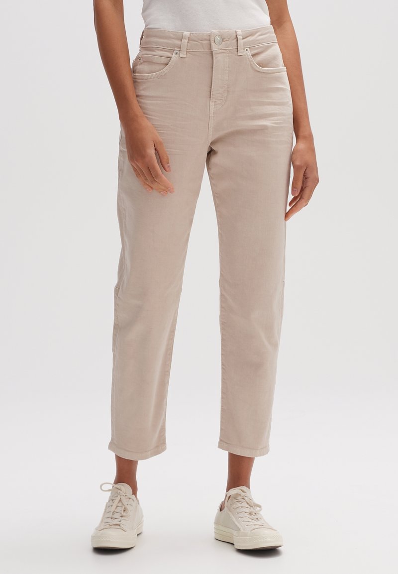 Pantalon beige clair à jambes droites en coton doux. Il possède des poches avant, une fermeture par bouton et des revers roulés. Porté avec des baskets blanches.