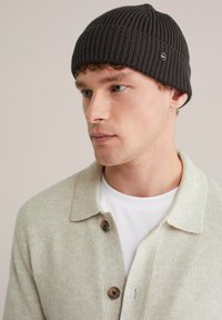 Căciulă tricotată de culoare gri închis, cu textură ribbed și un mic logo, purtată de o persoană îmbrăcată cu un cardigan bej deschis și o cămașă albă dedesubt.