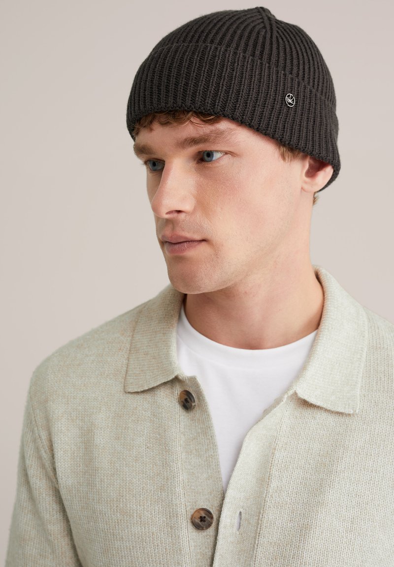 Căciulă tricotată de culoare gri închis, cu textură ribbed și un mic logo, purtată de o persoană îmbrăcată cu un cardigan bej deschis și o cămașă albă dedesubt.