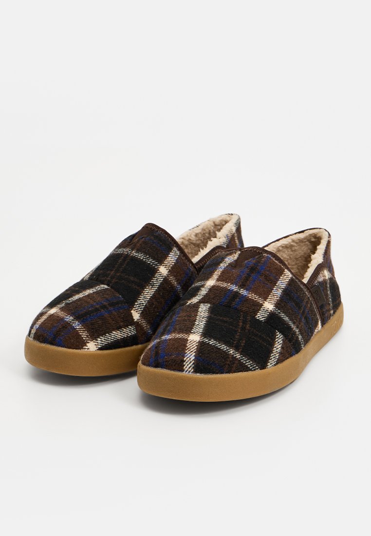 Pantuflas de cuadros marrones con un interior suave y forrado, presentando un diseño de tipo slip-on y una suela de goma color canela, acentuadas con rayas azules y blancas.