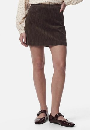 Femme portant une mini-jupe en velours côtelé marron, un chemisier à manches longues fleuri et des chaussures plates Mary Jane marron foncé, debout sur un fond blanc.