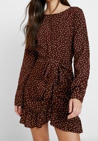Brun klänning med prickar, långa ärmar, rund halsringning, knytmidja och volangkant. Tillverkad av ett mjukt tyg med en slät textur.