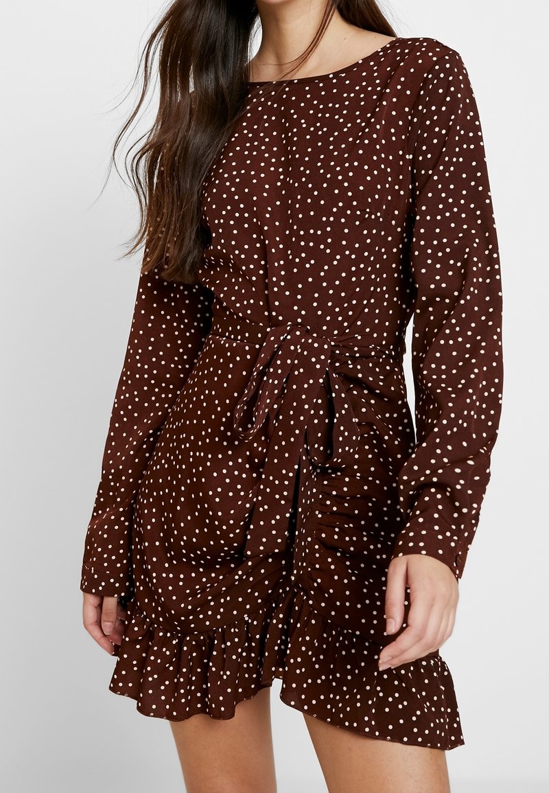 Brun klänning med prickar, långa ärmar, rund halsringning, knytmidja och volangkant. Tillverkad av ett mjukt tyg med en slät textur.