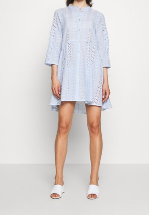 Mujer vestida con un vestido de encaje calado azul claro con mangas tres cuartos y botones frontales, combinado con sandalias mule blancas de tacón abierto.