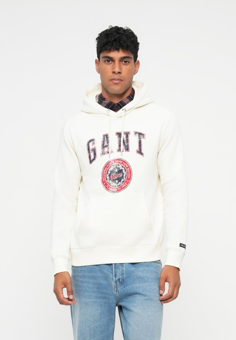 Felpa crema realizzata in morbido tessuto, caratterizzata da un grande logo "GANT" e una grafica circolare. Include una tasca frontale e un cappuccio con cordino.
