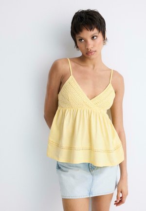 Młoda kobieta z krótkimi ciemnymi włosami, ubrana w żółty, bezrękawnik i jasnoniebieską, dżinsową mini spódnicę, stojąca na tle białej ściany.