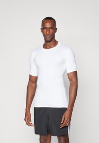 Hvit treningst-skjorte med korte ermer laget av et elastisk og glatt stoff, kombinert med sorte shorts. Enkel rund hals, kroppsnær passform.