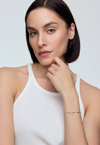 Złoty bransoletka z kolorowymi koralikami, wystawiona na dłoni opierającej się na brodzie. Modelka ma na sobie prosty biały top na ramiączkach. Neutralne tło.