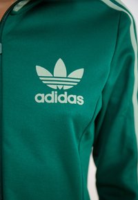 Türkiisi vettsjakko, mis on valmistatud tekstuuriga kangast, esiselt on valge Adidas'i logo ja kolme triibuga varrukatel.