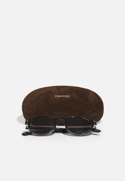 Schwarze Sonnenbrille mit ovalen Gläsern, die ein elegantes Design und sichtbares ‚TOM FORD‘-Branding aufweist, in einem braunen, weichen Wildlederetui platziert.