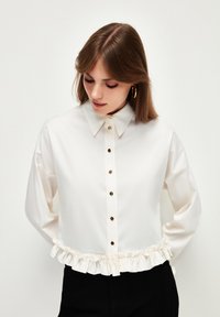 Camicia bianca con colletto a punta, bottoni dorati e orlo arricciato. Tessuto morbido; maniche lunghe con vestibilità morbida.