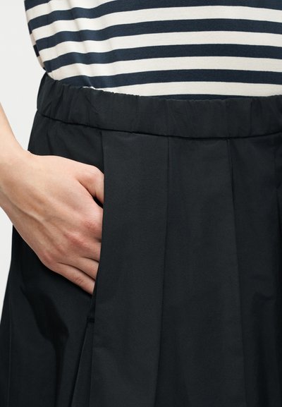 Χέρι που ακουμπά στην τσέπη μιας μαύρης πλισέ φούστας που φοριέται με ένα μαύρο και άσπρο ριγέ τοπ.