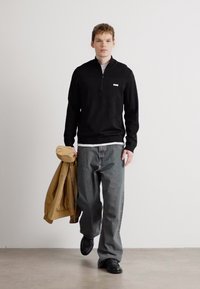 Μαύρο φούτερ με φερμουάρ, γκρι τζιν και μαύρα παπούτσια. Μοντέλο που κρατάει ένα μπεζ μπουφάν, στέκεται σε ένα δάπεδο ουδέτερου χρώματος μπροστά από φόντο απλής σχεδίασης.