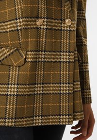 Blazer croisé de couleur marron terreux avec un motif en quadrillage. Présente des boutons dorés, un tissu texturé, et un design de col angulaire.