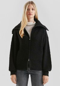 Zwarte fleecejack met een brede kraag, ritssluiting aan de voorkant en oversized mouwen. Heeft een gestructureerde stof en ribgebreide manchetten.