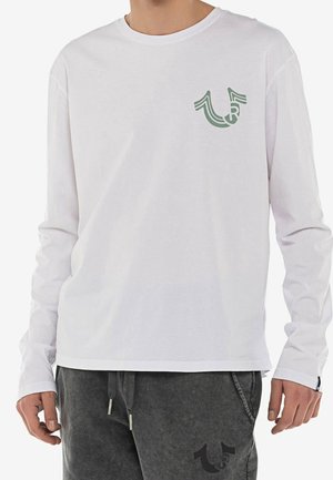 Homme portant une chemise blanche à manches longues avec un petit logo vert sur la poitrine et un pantalon gris foncé avec un logo assorti.