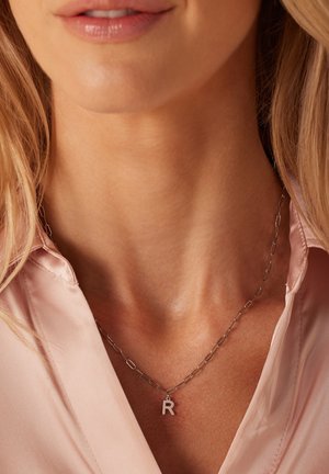 Collier en argent avec un design de maillons de chaîne et un pendentif en forme de lettre "R", porté sur une chemise rose pâle.