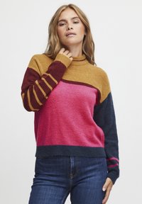 Pull multicolore avec des blocs jaunes, bordeaux, roses et bleu foncé. Texture douce, col montant et détails de manches rayées.