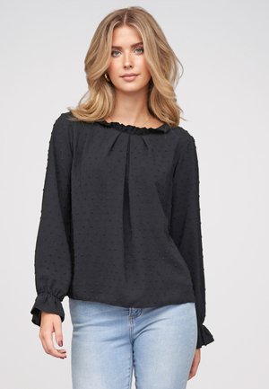 Femme blonde aux cheveux détachés portant un chemisier noir texturé à manches longues et un jean bleu clair sur un fond clair uni.