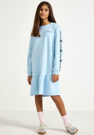 Młoda dziewczyna stoi, ubrana w jasno niebieską sukienkę z długim rękawem z czarnymi kokardkami na rękawach, różowe skarpetki i srebrne tenisówki na białym tle.