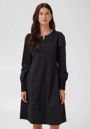 Femme aux longs cheveux bruns portant une robe noire à manches longues avec un col décoré et des poignets smockés, debout devant un fond gris.
