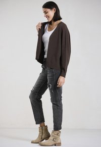 Brown oversized cardigan over a white tank top, paired with distressed black jeans and beige ankle boots with buckle accents.