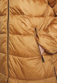 Doudoune de couleur brun doré chaud, présentant une texture matelassée, des poches latérales zippées et une finition lisse et brillante.