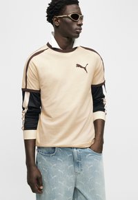 Νεαρός άνδρας με μπεζ φούτερ Puma με μαύρα και λευκά μανίκια, τζιν παντελόνι με ανοιχτό μπλε λογότυπο Puma και μπεζ γυαλιά ηλίου, κοιτάζει προς τα πλάγια.