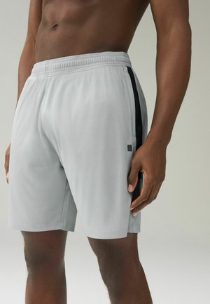 Man wearing light gray athletic shorts with black side stripes standing against neutral background.