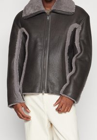 Brun læderjakke med grå shearling-detaljer, lynlåslukning og bred krave. Har en tekstureret yderside og blødt inderfor.