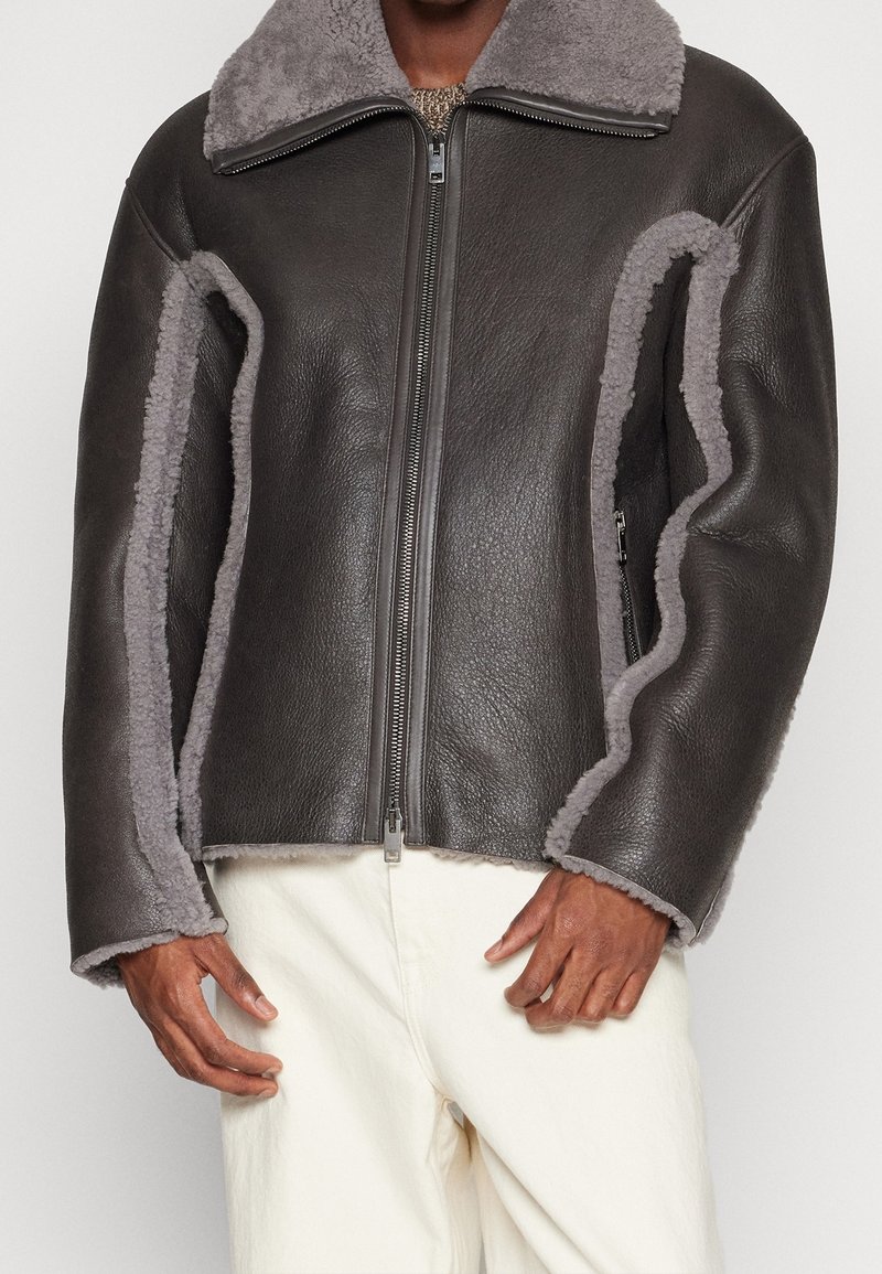 Brun læderjakke med grå shearling-detaljer, lynlåslukning og bred krave. Har en tekstureret yderside og blødt inderfor.