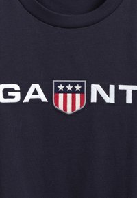 Námořnická modrá bavlněná tričko s bílými vyšitými písmeny "GANT", s logem ve štítu v červené, bílé a modré barvě, a třemi bílými hvězdami.