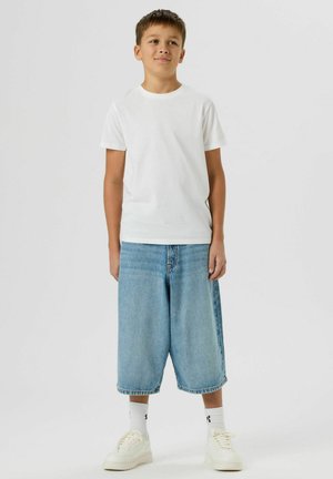 Ein Junge steht vor einem einfarbigen Hintergrund und trägt ein schlichtes weißes T-Shirt, lockere hellblaue Jeansshorts, weiße Socken und weiße Sneakers.