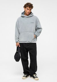 Lichtgrijze oversized hoodie met een voorzak en geborduurd logo, gecombineerd met zwarte baggy broek en zwarte sneakers met witte zolen.