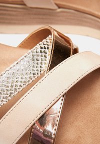 Sandales compensées beiges en daim lisse avec des accents métalliques et une sangle texturée, mettant en valeur un mélange de finitions et un design superposé.