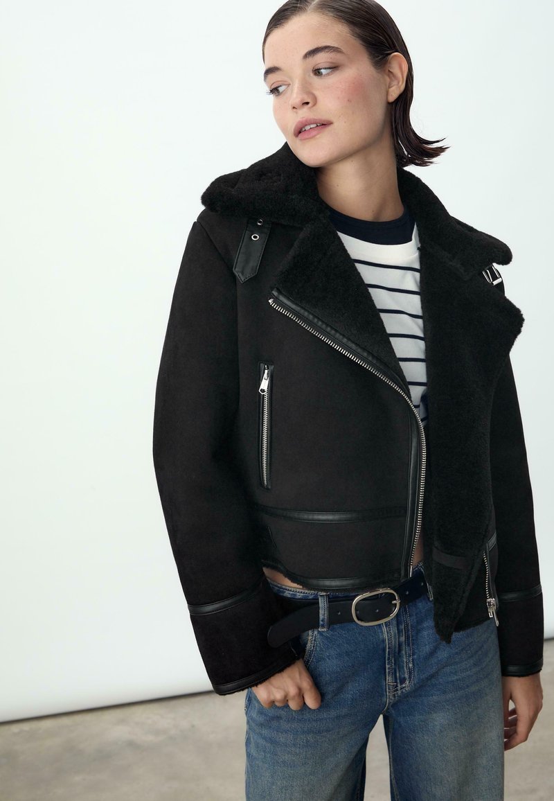 Czarna kurtka z owczej wełny z puszystym kołnierzem, zamykanymi na zamek kieszeniami, skórzanymi akcentami, krótki krój, noszona na w paski koszuli, zestawiona z niebieskimi dżinsami.