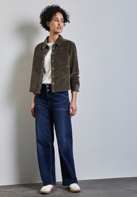 Chaqueta de pana marrón con botones, camisa blanca, vaqueros de pierna ancha azul oscuro y zapatillas ligeras. Diseño simple y texturizado con colores neutros.