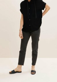 Camicia nera a maniche corte con bottoni e taschino sul petto, abbinata a jeans grigio scuro e sandali neri piatti. Sfondo con superficie piatta.