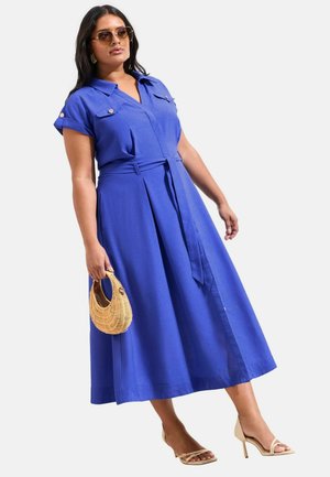 Frau in einem blauen Midikleid mit Gürtel, die eine Korbtasche hält, beigefarbene High Heels trägt und eine Sonnenbrille aufhat.