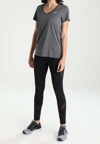 Grå v-ringad träningsskjorta, åtsittande svarta leggings med nätpaneler, och grå sneakers med mörka snören; avslappnad sportoutfit.