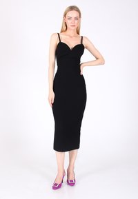 Abito midi aderente senza maniche di colore nero con scollatura a cuore, abbinato a scarpe con tacco alto viola dotate di un'applicazione quadrata.