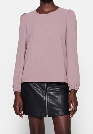 Blusa lilla con maniche lunghe e arricciate e scollatura rotonda, abbinata a una minigonna in pelle nera con zip centrale.
