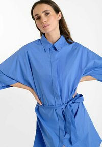 Vestido camisero azul de gran tamaño, hecho de tela ligera, con un cuello tipo polo, mangas amplias y un cinturón atado en la cintura para dar forma.
