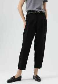 Pantalon noir sur mesure taille haute, avec plis sur le devant et deux poches latérales, associé à un haut gris à manches courtes et des mocassins noirs.